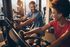 Vibrant gym scene with people enjoying a cheerful workout on stationary bikes, surrounded by large windows and natural light.
