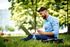 Man enjoying a relaxing work setup outdoors with a laptop and a sandwich, surrounded by green trees and bright sunlight.