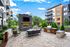 Chic outdoor lounge area with modern fireplace, cozy seating, and BBQ grill set against stylish apartment buildings and lush greenery.