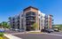 Modern apartment complex with spacious balconies, surrounded by lush greenery and ample parking spaces under a clear blue sky.