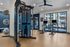 Modern fitness center with state-of-the-art gym equipment, including a weight machine and stationary bicycles, boasting floor-to-ceiling windows with a view of the apartment complex courtyard.