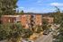 Charming brick apartment building nestled in a lush, tree-lined neighborhood with a serene street and distant forested hills.