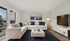 Modern living room with stylish white sectional sofa, elegant blue and gray accents, large TV, and floor-to-ceiling windows showcasing a scenic sunset view.