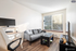 Bright, modern living room with sleek grey sofa, large window with natural light, and efficient home office setup on wood flooring.