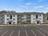 Modern three-story apartment building with elegant white facade and ample parking space, surrounded by lush greenery and clear blue skies.
