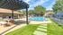 Green grass, brick and cement picnic table under a gazebo in the forground.  Pool with lounge chairs, trees in background.