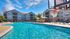Another community pool photo showing a basketball hoop, hanging over the water, lounge chairs, and water volleyball net in the distance.  Apartment buildings and blue sky fill the background.