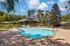 Community pool with clubhouse in background. Pool is shaded by tree in the foreground and surrounded by lounge chairs, tables, and umbrellas.