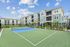 Modern apartment complex with a tennis court, featuring stylish architecture and lush landscaping under a bright blue sky.
