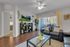 Bright living room with contemporary decor, featuring a sleek black sofa, glass coffee table, and entertainment unit with a scenic view on the TV. Sunlight pours in through large windows, enhancing the welcoming atmosphere.