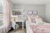 Cozy bedroom with elegant decor, featuring soft pink accents, plush pillows, and a stylish workspace illuminated by natural light.