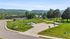 Scenic view of lush green parkland with a paved walkway, surrounded by mature trees and rolling hills under a clear blue sky.