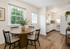 Charming dining area with a round wooden table and elegant chairs, connected to a modern kitchen with sleek white cabinets and large windows offering abundant natural light.