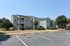 Charming apartment complex exterior with balconies and well-maintained landscaping, featuring ample parking spaces under a clear blue sky.