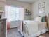 Boise, ID, Apartments With In-Unit Washer and Dryer - Kensington - Cozy Bedroom With White Metal Bed Frame, Soft Bedding, Large Window With Pink Curtains, Dresser, and Wall Shelves.