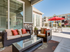 Two-Bedroom Apartments in Meridian, ID - Regency at River Valley - Outdoor Patio Furnished with Rattan-Made Cushion Sofa, Armchair, and Glass Top Coffee Table, and a Large Rug, Fronting the Clubhouse Facade with Oversized Windows, and Overlooking the Pool Area.