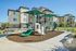 Modern apartment complex with a vibrant playground featuring slides and climbing structures, surrounded by beautiful landscaping and a paved pathway.