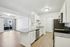 Modern apartment kitchen with sleek white cabinets, granite countertops, and stainless steel appliances, open to a bright living area with wood flooring and large sliding glass doors.