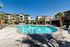 Luxurious apartment complex featuring a sparkling pool, surrounded by lush palm trees and modern architecture under a clear blue sky.