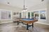 Contemporary apartment recreation room with elegant herringbone wood flooring, featuring a stylish pool table under a chic chandelier, surrounded by bright windows with scenic views.