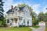 Charming two-story white house with inviting front porch, nestled in a lush green setting with a paved driveway leading to a spacious garage.