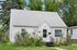Charming white cottage-style home with lush flower garden and inviting front steps, surrounded by greenery and trees.