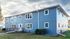 Charming two-story blue apartment building with symmetrical windows, surrounded by a lush green lawn and set against a cloudy sky.