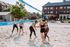 Enthusiastic group enjoying a sunny day playing beach volleyball on a sandy court, surrounded by lush greenery and modern brick buildings.