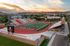 Stunning aerial view of a modern stadium with vibrant red seating and lush green field, set against a breathtaking sunset and scenic landscape.