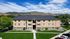 Charming three-story apartment building with scenic mountain backdrop, surrounded by lush green lawns and clear blue sky.