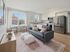 Bright and modern open-concept living space featuring a sleek kitchen with stainless steel appliances, a stylish grey sofa, and a cozy dining area with large window views.