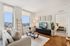 Bright and modern living room with large windows offering city views, featuring a cozy setup with a plush couch, stylish decor, and a sleek TV unit, seamlessly connecting to a comfortable bedroom.