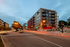 Modern urban apartment building with vibrant red accents and spacious balconies, nestled in a lively cityscape at dusk.
