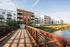 Modern riverside apartments with spacious balconies, surrounded by lush greenery, connected by a charming wooden bridge, offering serene water views.