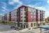 Modern five-story apartment building with red and beige exterior, featuring balconies and street-level shops.