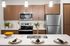 Queen Creek, AZ Apartments - Acero Harvest Station - View Of The Beautiful Kitchen From Behind The Kitchen Island Bar Area With Three Plates Set, The Kitchen Has Stainless Steel Appliances And Modern Style Wood Cabinetry