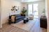Modern living room with chic decor, featuring a plush gray sofa adorned with accent pillows, stylish abstract wall art, and a cozy armchair by a large window overlooking green trees.