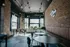 Chic industrial-style dining area with large windows, exposed brick walls, and contemporary furniture, featuring pendant lighting and lush greenery.