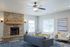 Inviting living room with a cozy stone fireplace, elegant ceiling fan, and contemporary furnishings, accented by large windows and tasteful decor.