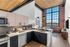 Riverwalk Lofts | Modern loft apartment with high ceilings and large windows. Features a kitchen with light wood and black cabinets, gray backsplash, and stainless steel appliances. Brick accent wall and a living area with a light sofa and a large TV.| Loft apartments for rent lawrence ma
