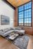 Riverwalk lofts |  A modern living room with exposed brick walls and a large window showing a cityscape. It features a gray sectional sofa, a metallic coffee table with decor, and a gray rug. A framed artwork hangs on the wall, and the ceiling is wooden.| Apartments for rent lawrence ma