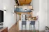 Riverwalk Lofts | Modern loft apartment with a high ceiling and exposed wooden beams. The kitchen features sleek white cabinets and stainless steel appliances, with a breakfast bar and two gray stools. Above, a frosted glass partition hints at an upper level. | Studio Apartments lawrence ma