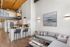 Riverwalk Lofts | Modern loft apartment with a high ceiling and exposed wooden beams. The kitchen features sleek white cabinets and stainless steel appliances, with a breakfast bar and two gray stools. Above, a frosted glass partition hints at an upper level. | Studio Apartments lawrence ma