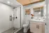 riverwalk lofts | Modern bathroom with a glass-enclosed shower, white tiles, a wooden ceiling, and a light wood vanity. A lit mirror is above the sink. A colorful abstract painting and a vase with flowers are on the vanity. | luxury apartments lawrence ma