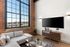 riverwalk lofts | A modern living room with a large loft window, exposing city views. It features exposed brick walls, a gray sectional sofa, a sleek wooden TV console, and a wall-mounted TV. A glass coffee table and a small potted plant complete the decor. | apartments for rent near boston