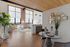 Riverwalk Lofts | A modern, open-concept loft apartment with large windows, exposed brick, and wooden ceilings. The room features a dining area with a glass table, a kitchen with pendant lights, and a cozy seating area. A plant and geometric wall art add decorative touches.| Loft apartments lawrence ma