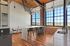 Riverwalk Lofts | A modern kitchen and dining area with high ceilings, featuring a large island with black stools, wooden floors, and industrial-style brick walls. Large windows let in natural light, and a small round table with two chairs is in the corner. | luxury apartments for rent lawrence ma