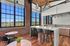 Riverwalk Lofts | Modern industrial-style kitchen with exposed brick walls, large windows offering a scenic view. Features a sleek marble island with black bar stools, hanging spherical lights, and two small round tables with chairs. | luxury apartments for rent lawrence ma
