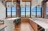 Riverwalk Lofts | Modern kitchen interior with large industrial-style windows, brick walls, and wooden flooring. Features a long marble island with bar stools, a countertop with a sink, and geometric hanging lights. The view through the windows shows a cityscape. | luxury apartments for rent lawrence ma