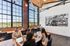 Riverwalk Lofts | A modern office with five people working at a long table using laptops. Large windows allow natural light, showcasing a scenic view outside. A black and white industrial-themed artwork decorates the brick and white walls. | loft apartments near boston